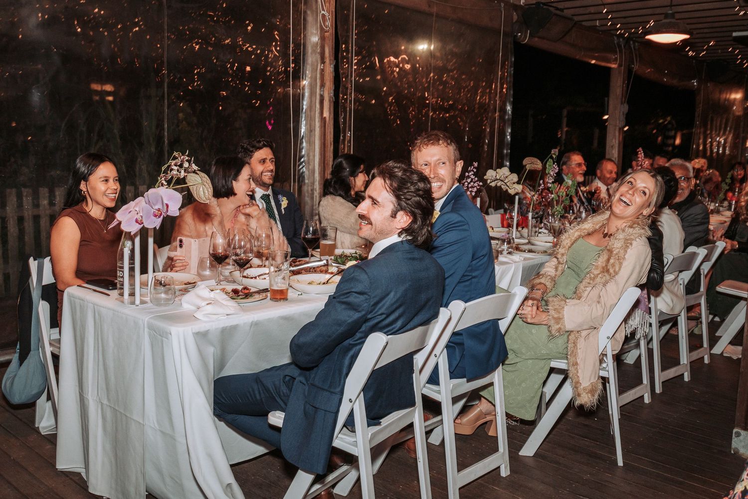 Wedding guests share laughter and conversation at elegantly decorated reception tables in rustic indoor venue.