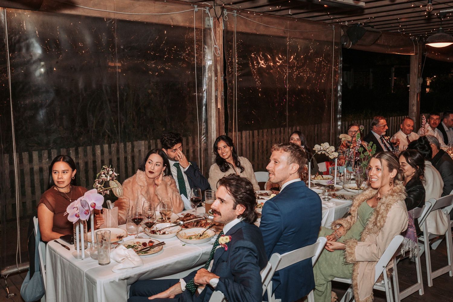 Wedding guests enjoy dinner and conversation at long tables during an intimate indoor reception celebration.