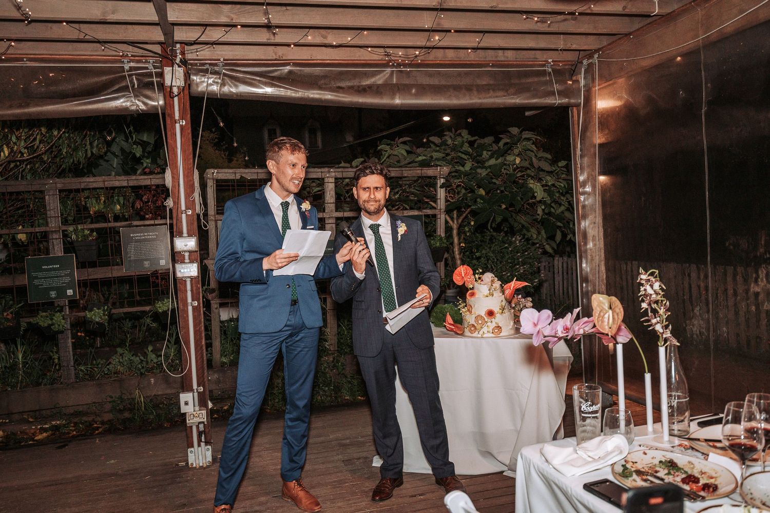 Two people in suits giving speeches during an elegant outdoor reception with decorated dining tables in a rustic venue.