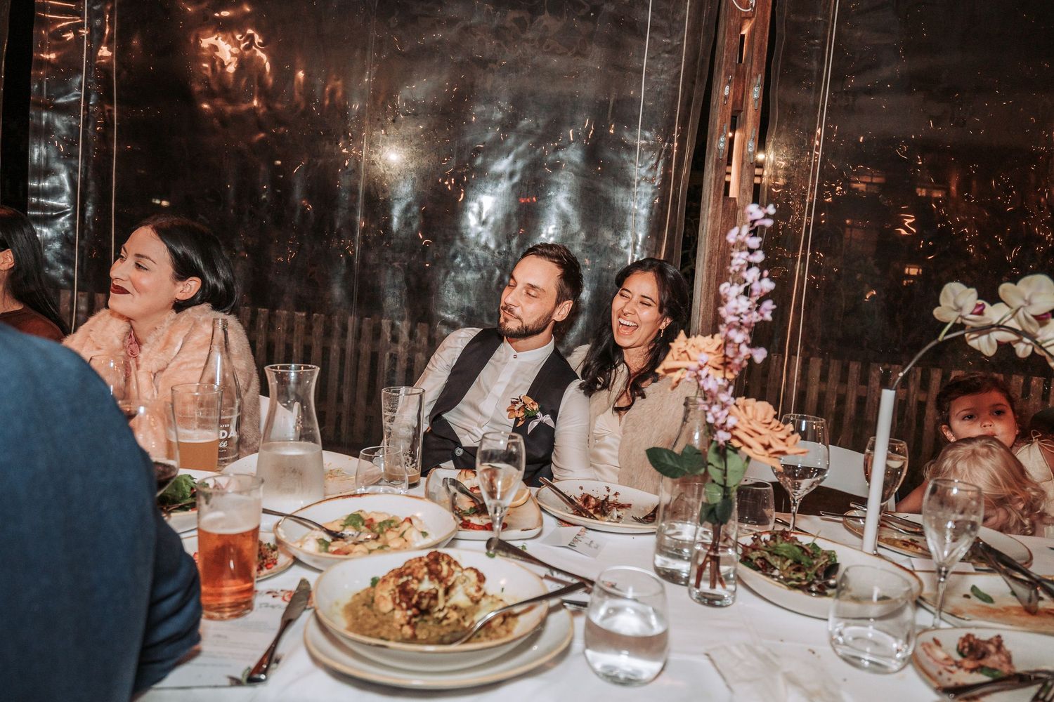 Candlelit dinner celebration at an elegant glass-enclosed venue with guests enjoying food and drinks at a decorated table.