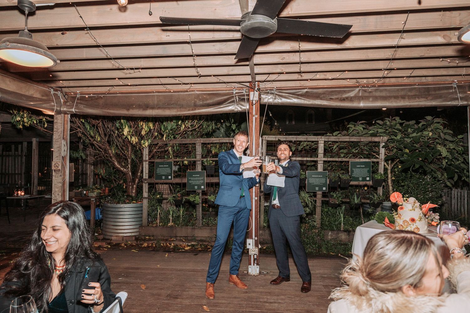 People celebrating under a rustic covered patio with string lights and wooden beams at an outdoor event venue.