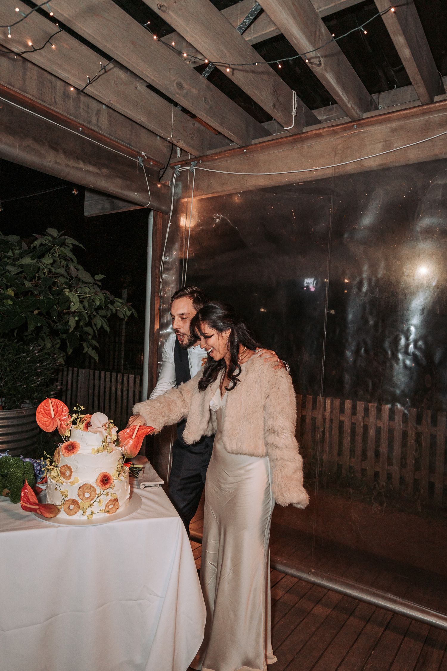 Couple cuts their elegantly decorated wedding cake with floral accents on a covered outdoor deck at night.