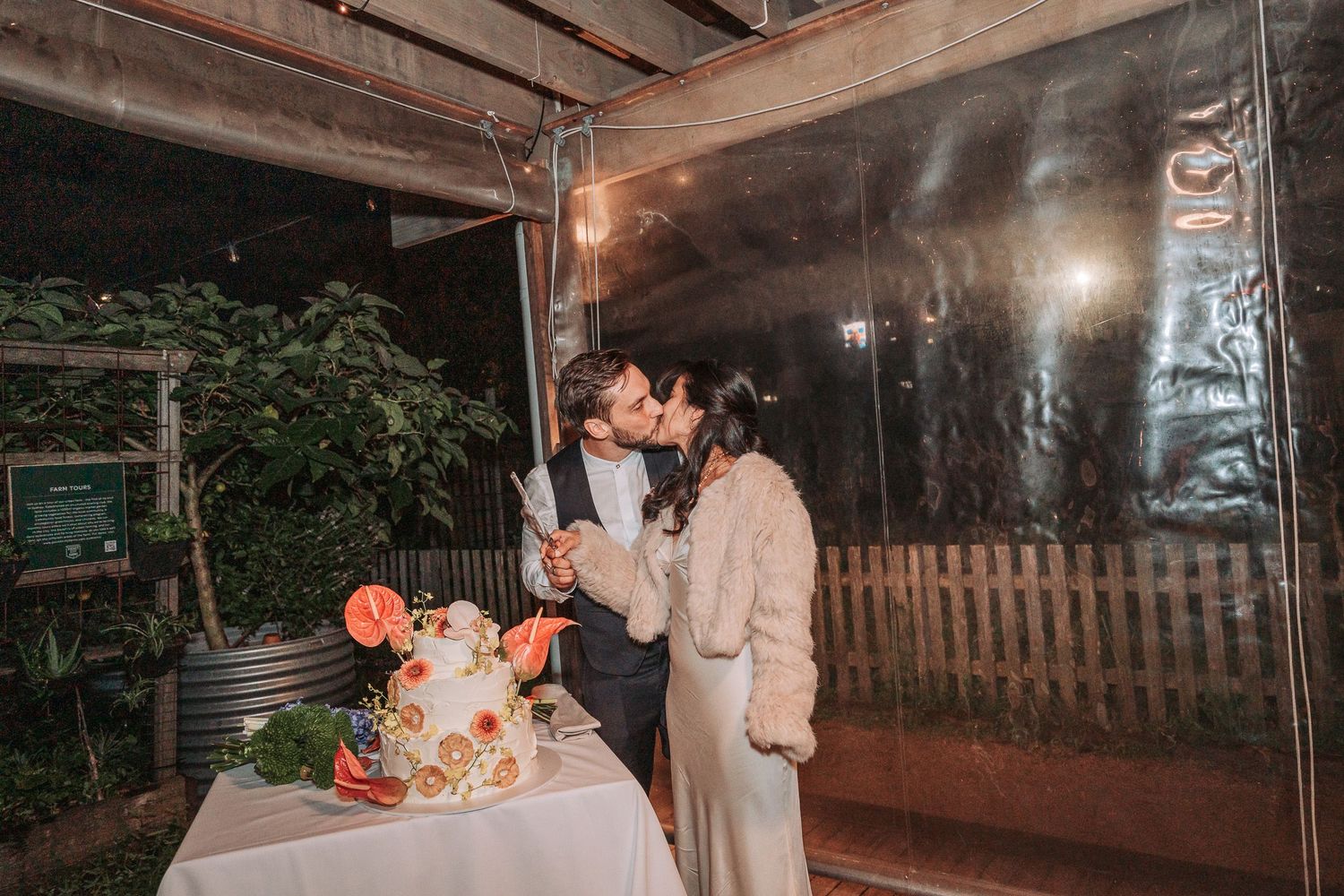 A romantic couple sharing a moment at an outdoor reception with a decorated table and glowing lights at night.