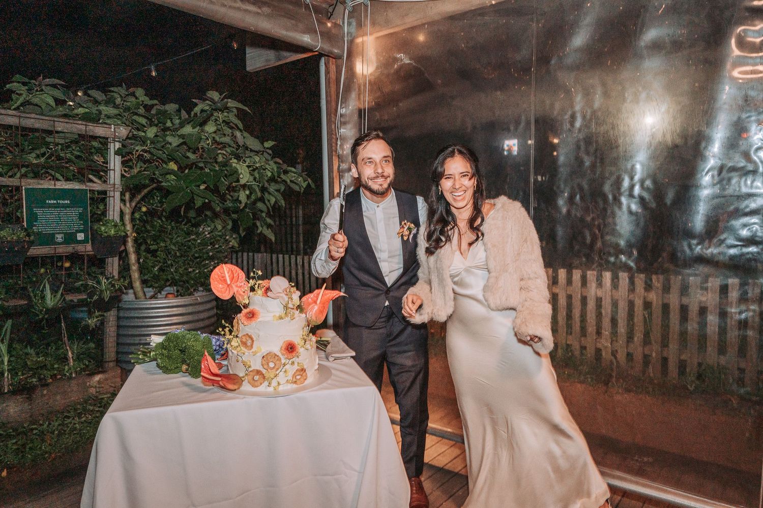 A couple poses next to their rustic wedding cake at an outdoor reception with garden decor and string lights.