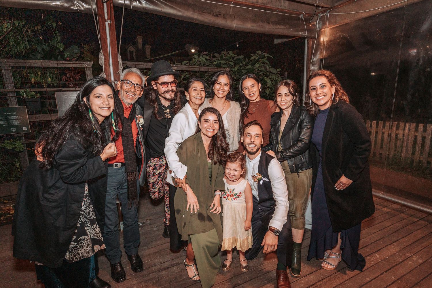 A group of people smiling together on a wooden deck at night with outdoor lighting.
