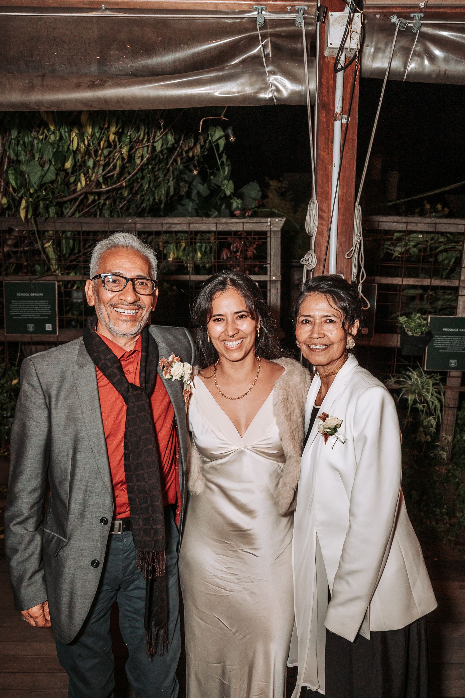 Three people stand together at a wedding celebration under a tent canopy at night.