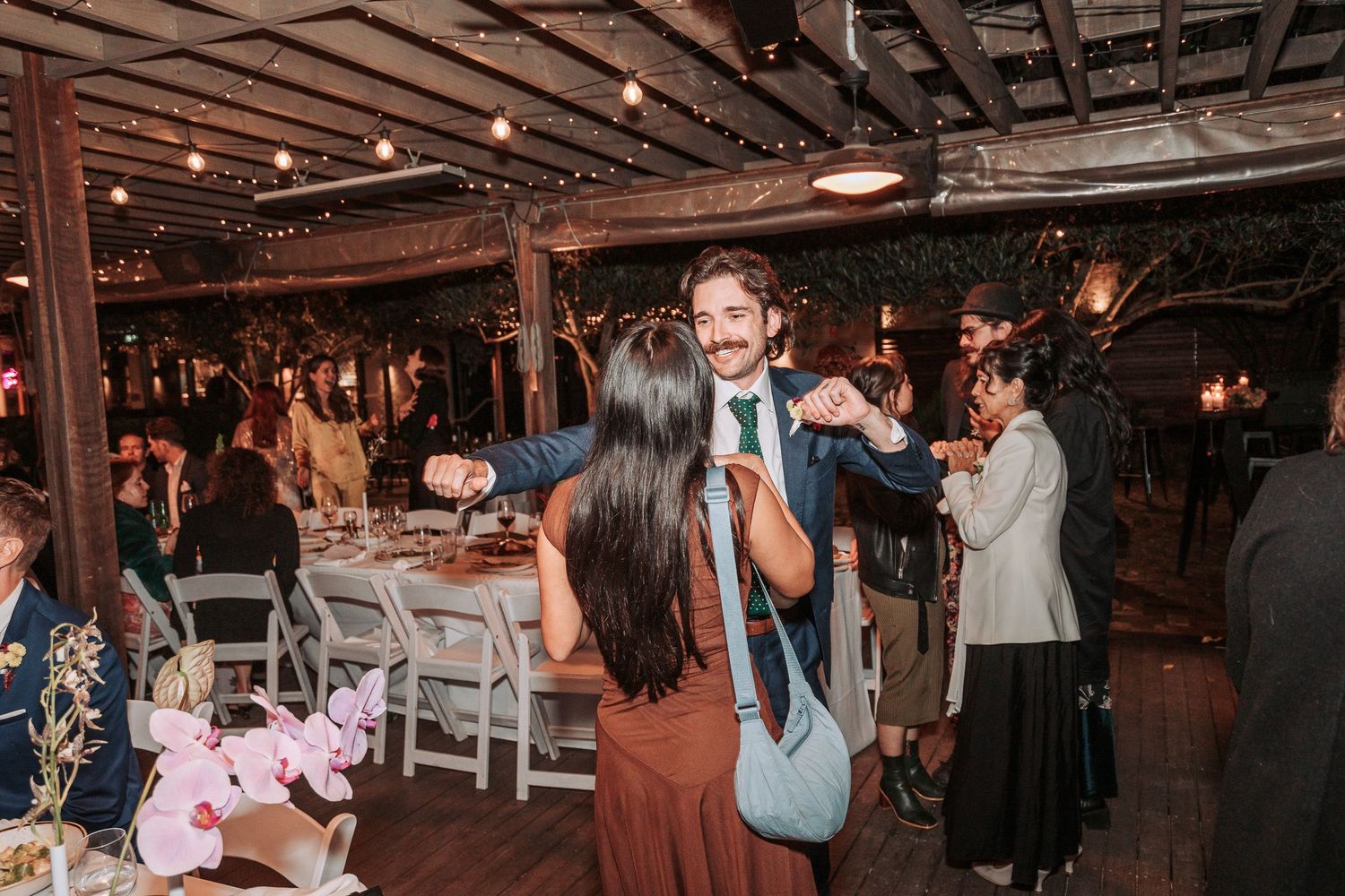People dancing at an indoor wedding reception with exposed ceiling beams and string lights creating a rustic atmosphere.