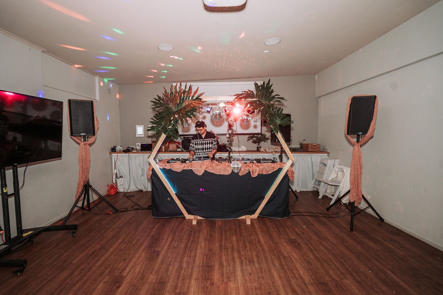 A professional DJ setup with speakers and equipment on a table with black cloth and decorative palm plants in an indoor venue.