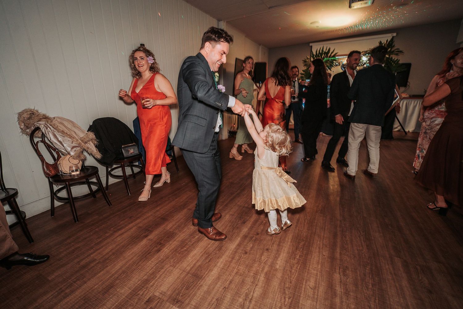 People dancing on a hardwood floor at a wedding reception with guests wearing formal attire in red dresses and suits.