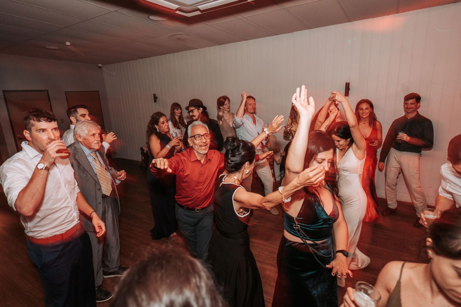 Party guests dancing enthusiastically on a wooden dance floor at an indoor celebration with warm lighting.