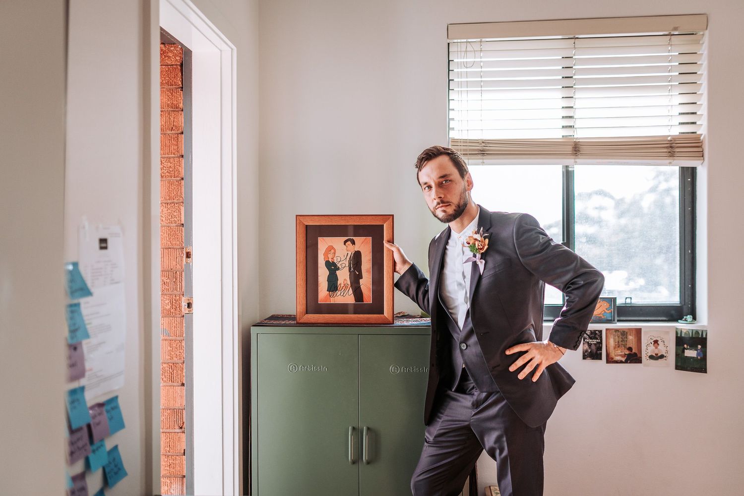A person in business attire poses casually against a green cabinet near a window in an office setting.