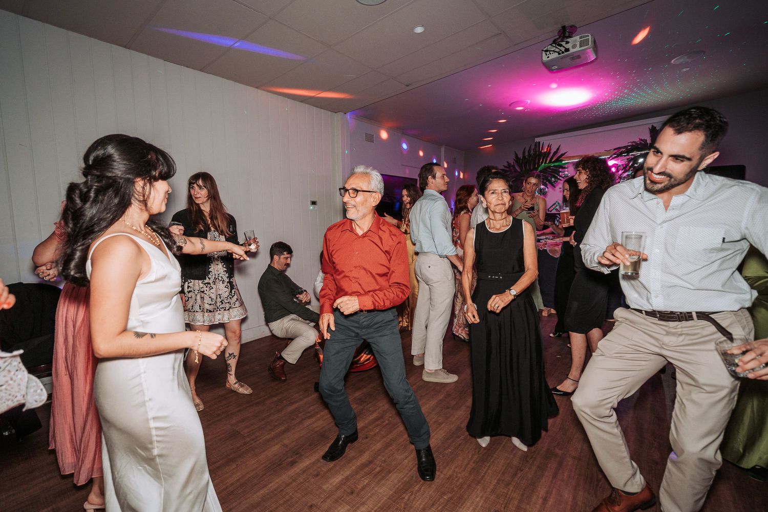 Party guests dance and socialize on a wooden dance floor under colorful pink and purple lighting at an indoor event.