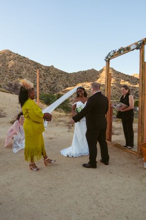 A desert wedding ceremony takes place under a wooden arch with mountains in the background at sunset.
