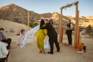 Desert wedding ceremony takes place at golden hour under wooden arch frame with mountains in background.