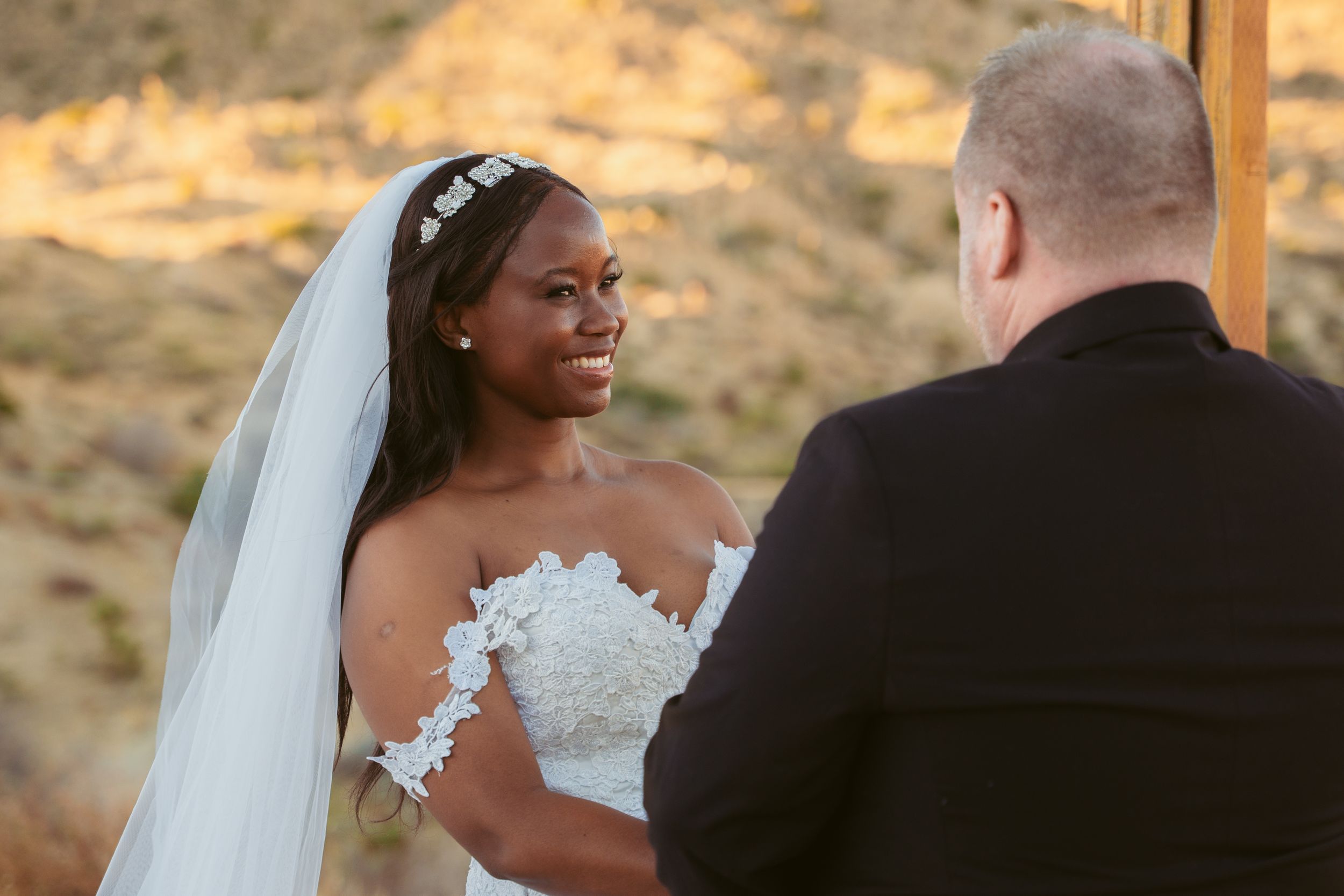 Couple exchanges vows during outdoor desert wedding ceremony beneath wooden arch at sunset.