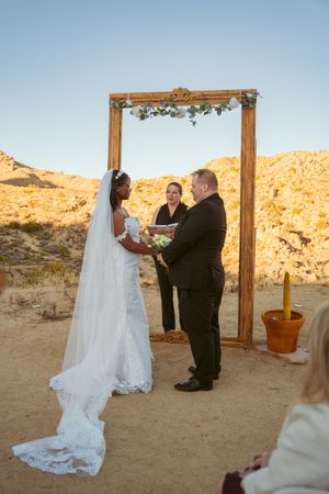 A wedding ceremony takes place in a desert landscape with a rustic wooden arch decorated with greenery at sunset.