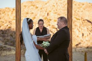 A couple exchanges vows during an outdoor desert wedding ceremony with an officiant under a wooden arch.