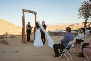 A wedding ceremony takes place at sunset in a desert setting with a wooden arch and seated guests.