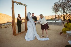 A wedding ceremony takes place at sunset in a desert setting with a wooden arch frame and attendees gathered around.