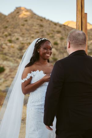 A joyful bride in a white lace dress and veil shares a candid moment during an outdoor desert wedding ceremony.