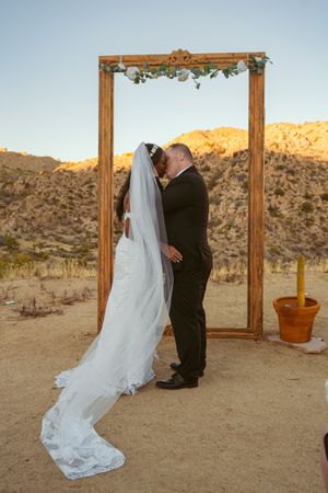 Newlyweds share first kiss under rustic wooden arch decorated with greenery in desert setting.