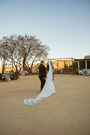 A couple stands in a desert landscape during golden hour with rustic decor and trees silhouetted against the sky.