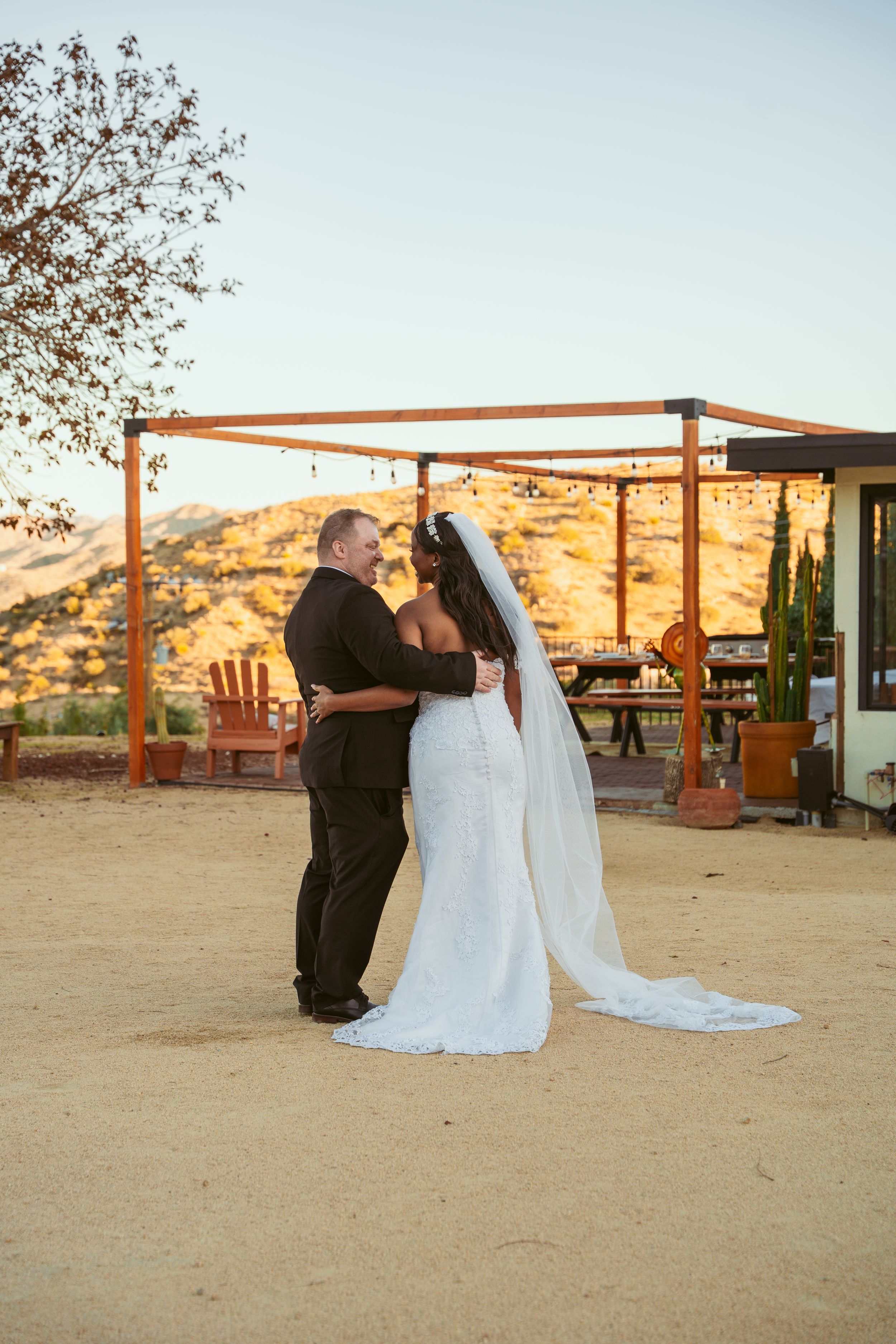 Newlyweds share an intimate moment at sunset under a wooden pergola in a desert landscape setting.