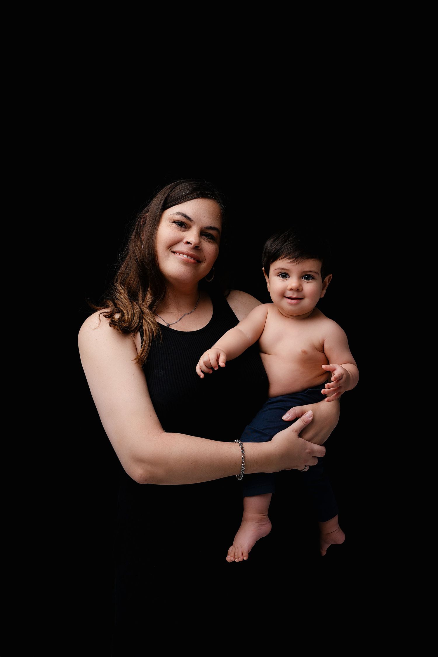 A woman in a black tank top holds a happy shirtless baby with dark hair against a black background.
