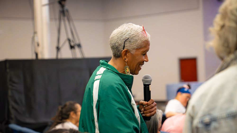 An elderly speaker in a green shirt addresses an audience with a microphone during an indoor event.