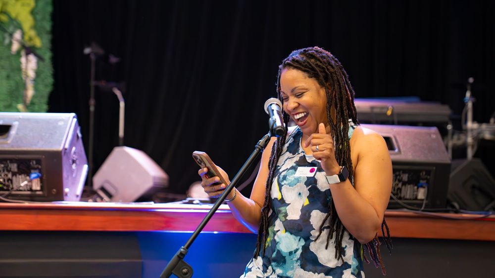 A performer in a floral dress sings enthusiastically on stage with audio equipment in the background.
