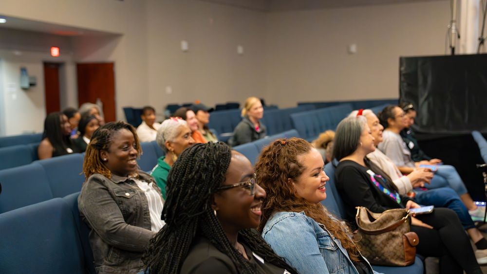 Engaged audience members smile and react while seated in blue chairs during a presentation.