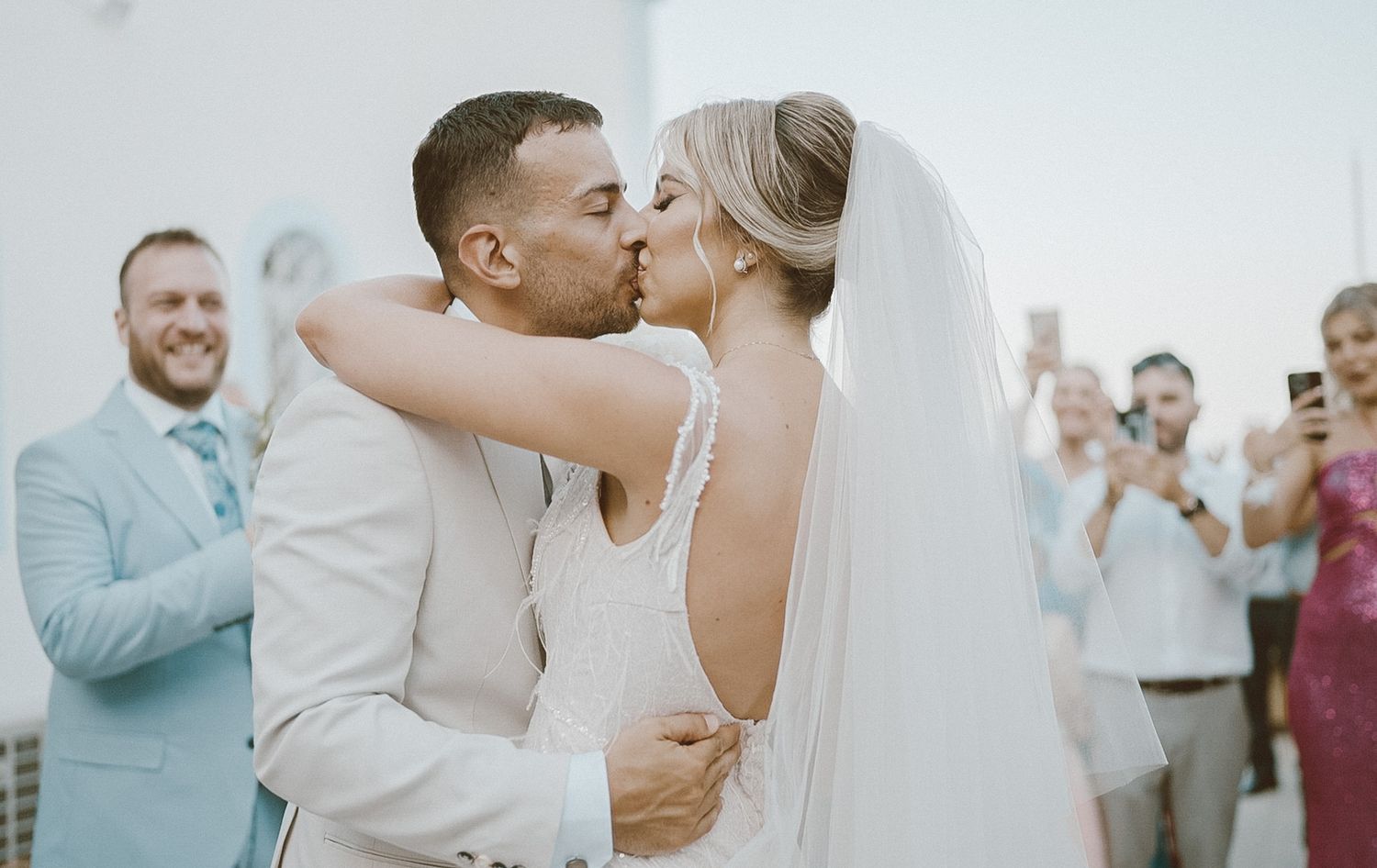 A romantic outdoor wedding ceremony captured in soft light as a couple shares their first kiss while guests applaud.