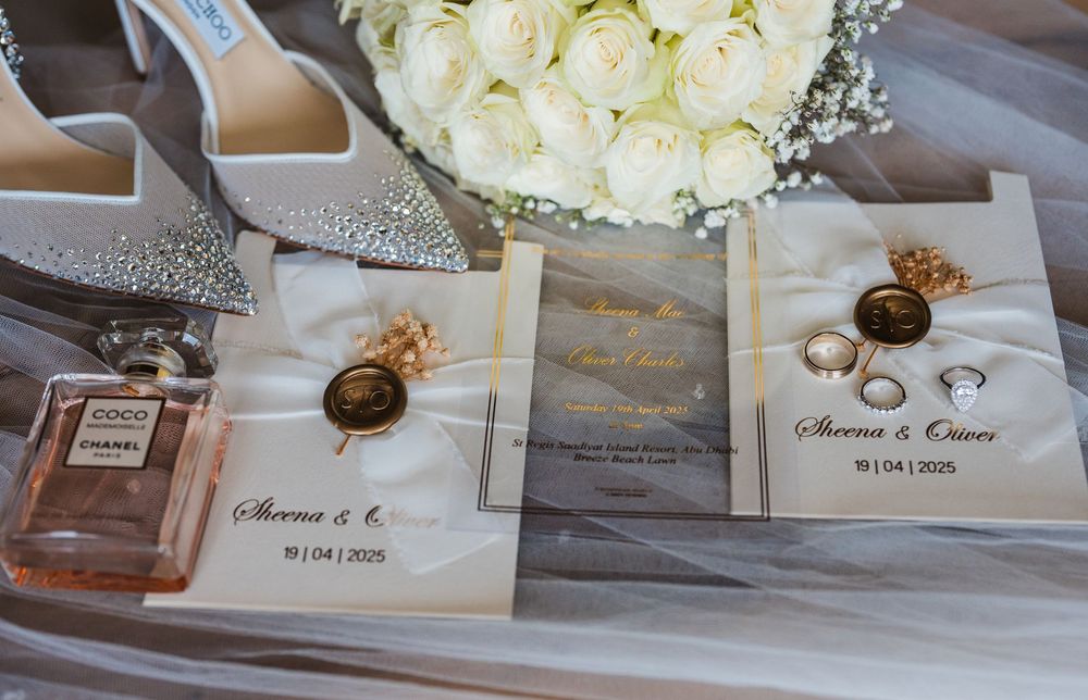 Wedding details including rings on invitations, perfume, silver shoes and white rose bouquet arranged on gray surface.
