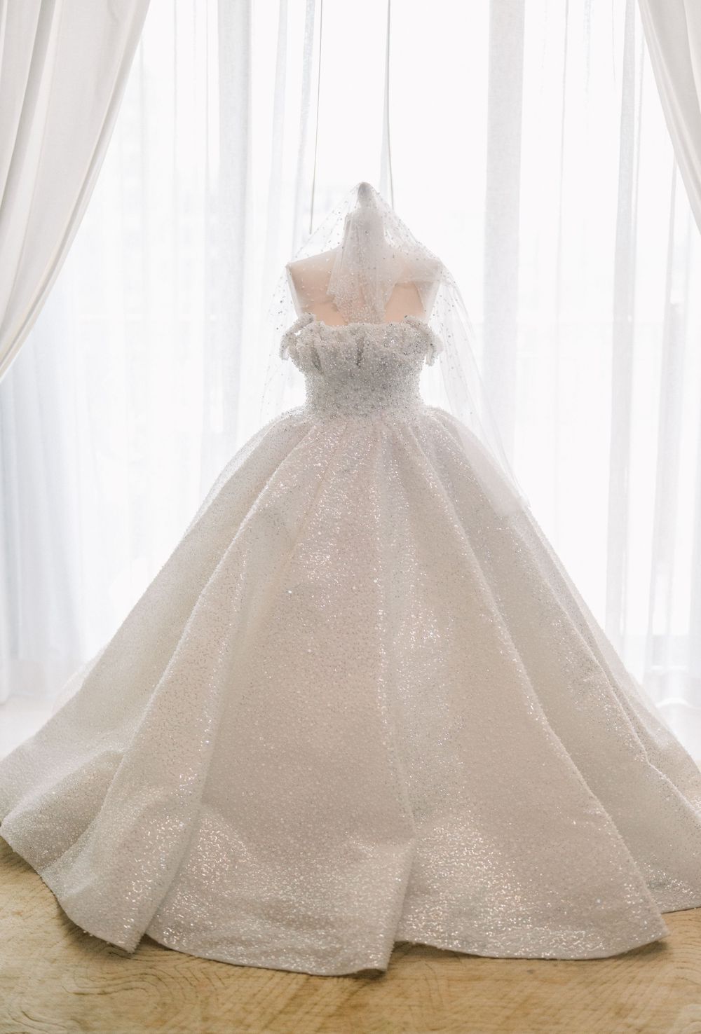 A luxurious white ball gown wedding dress with strapless bodice and full skirt displayed against sheer curtains.