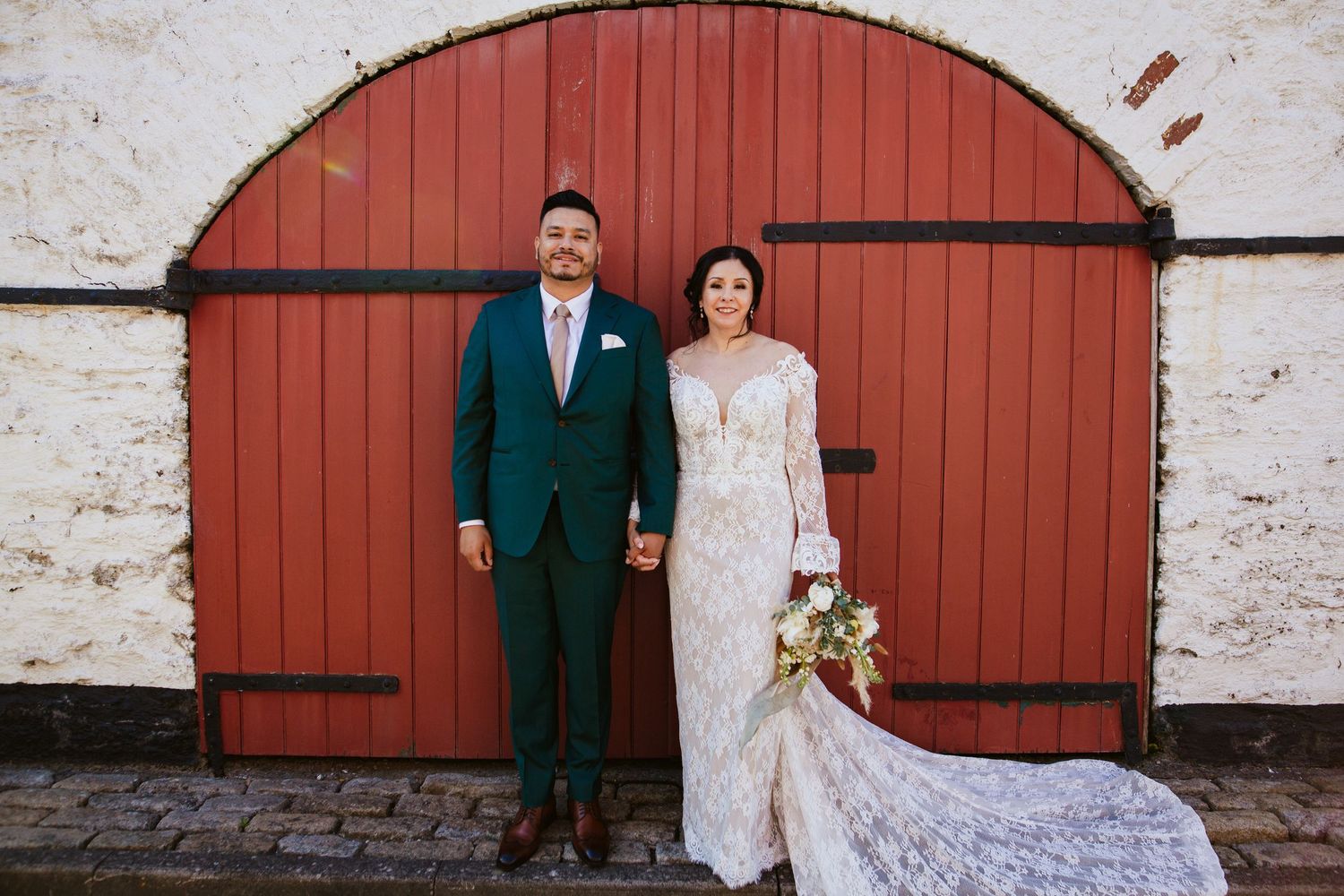 A couple poses in front of a rustic red barn door, with the bride wearing a flowing lace gown and holding a bouquet.
