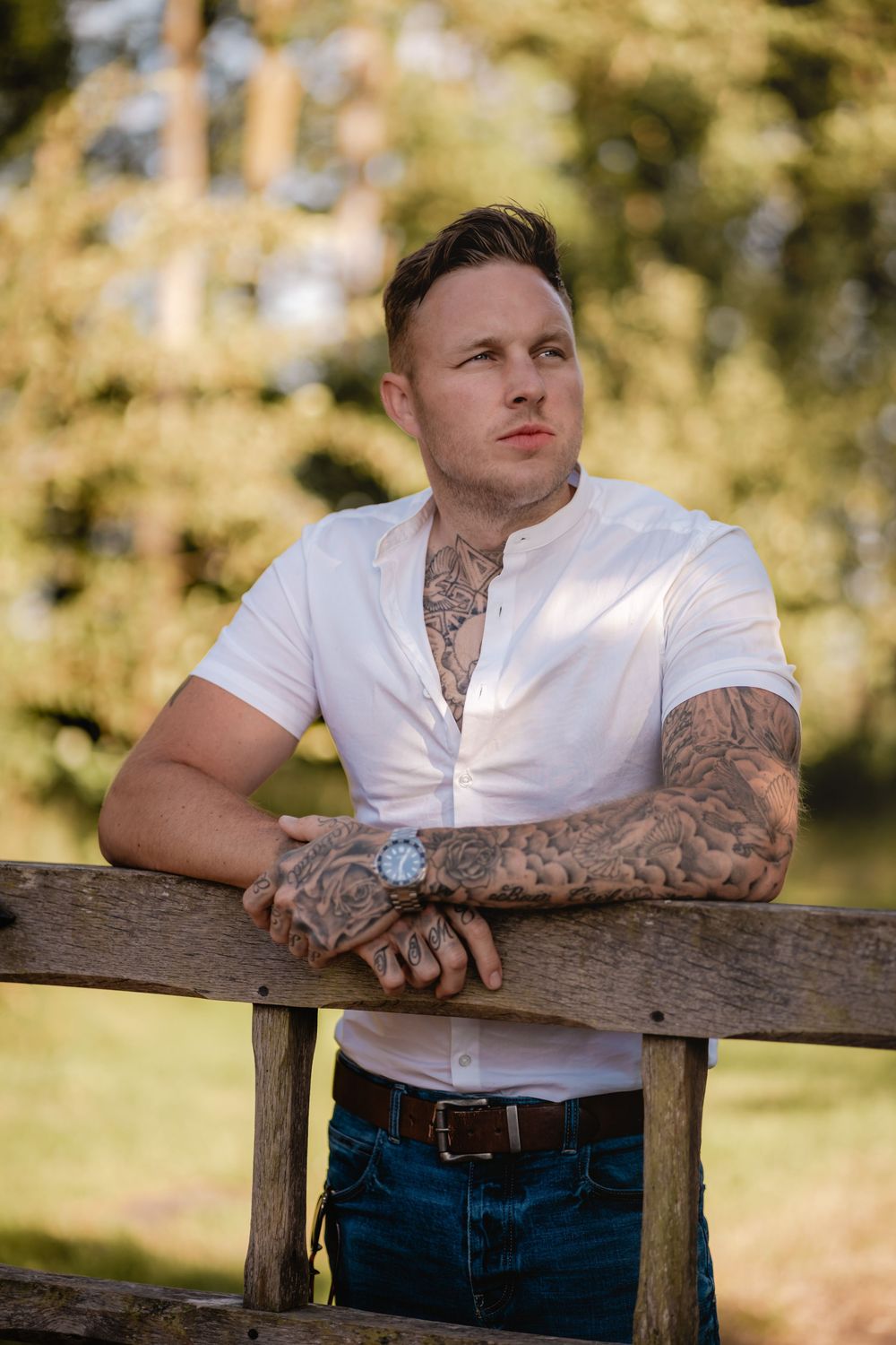 Portrait of person wearing white shirt leaning on wooden fence with visible arm tattoos.