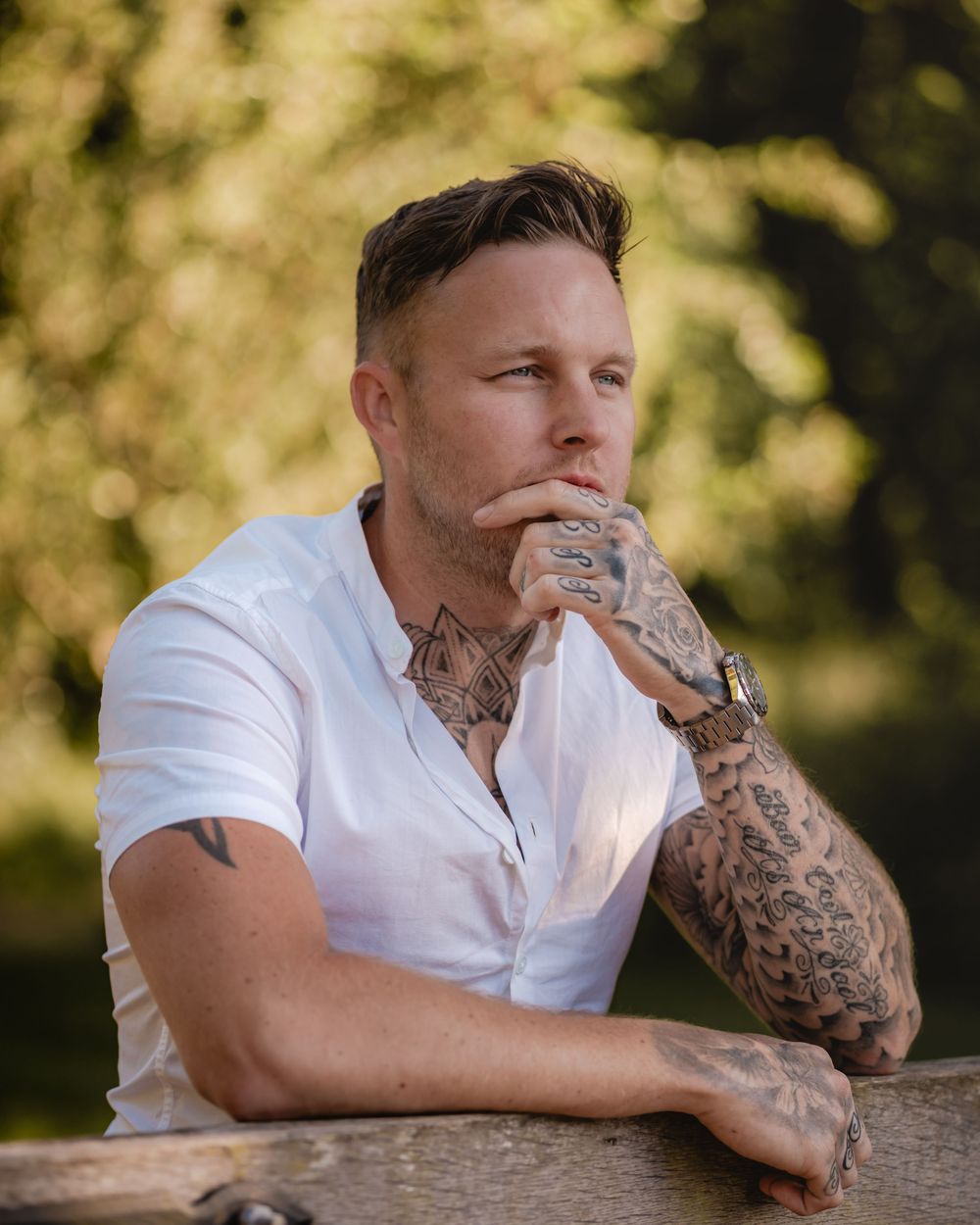 Portrait showing person in white button-up shirt with tattooed arm resting on wooden railing.