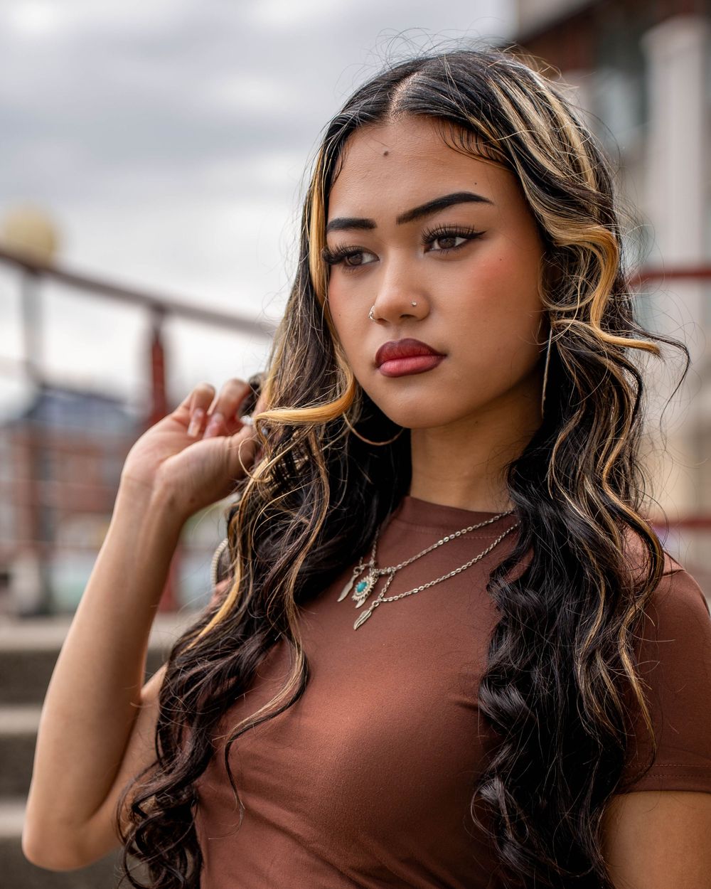 Fashion portrait featuring stylish jewelry and brown top against urban backdrop.