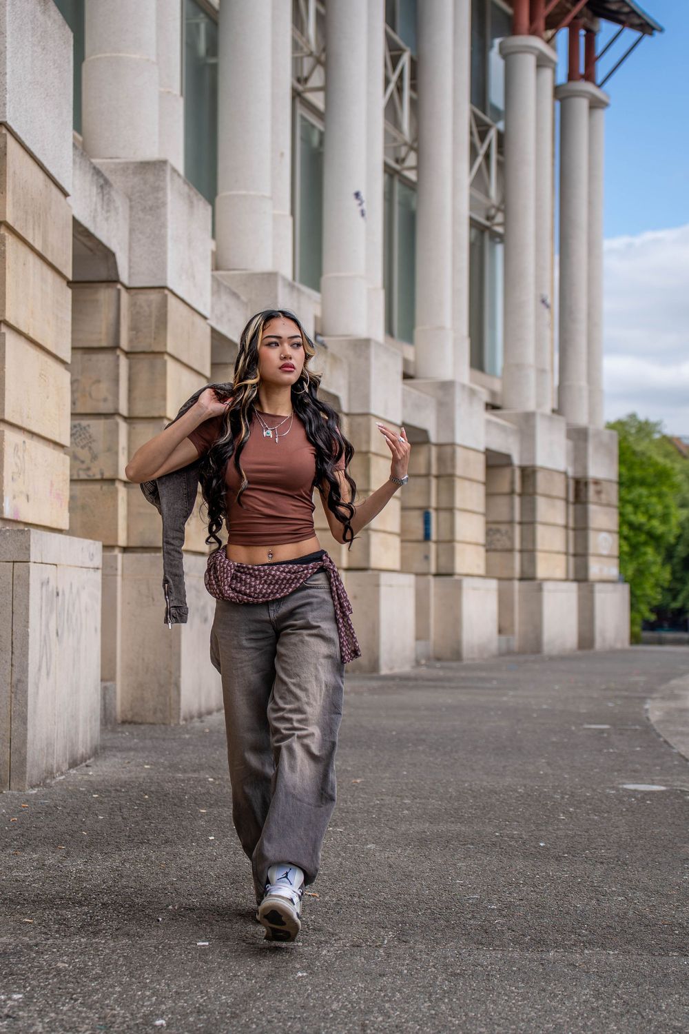 Urban fashion photography of a model in a brown crop top and cargo pants walking along a columned building exterior.