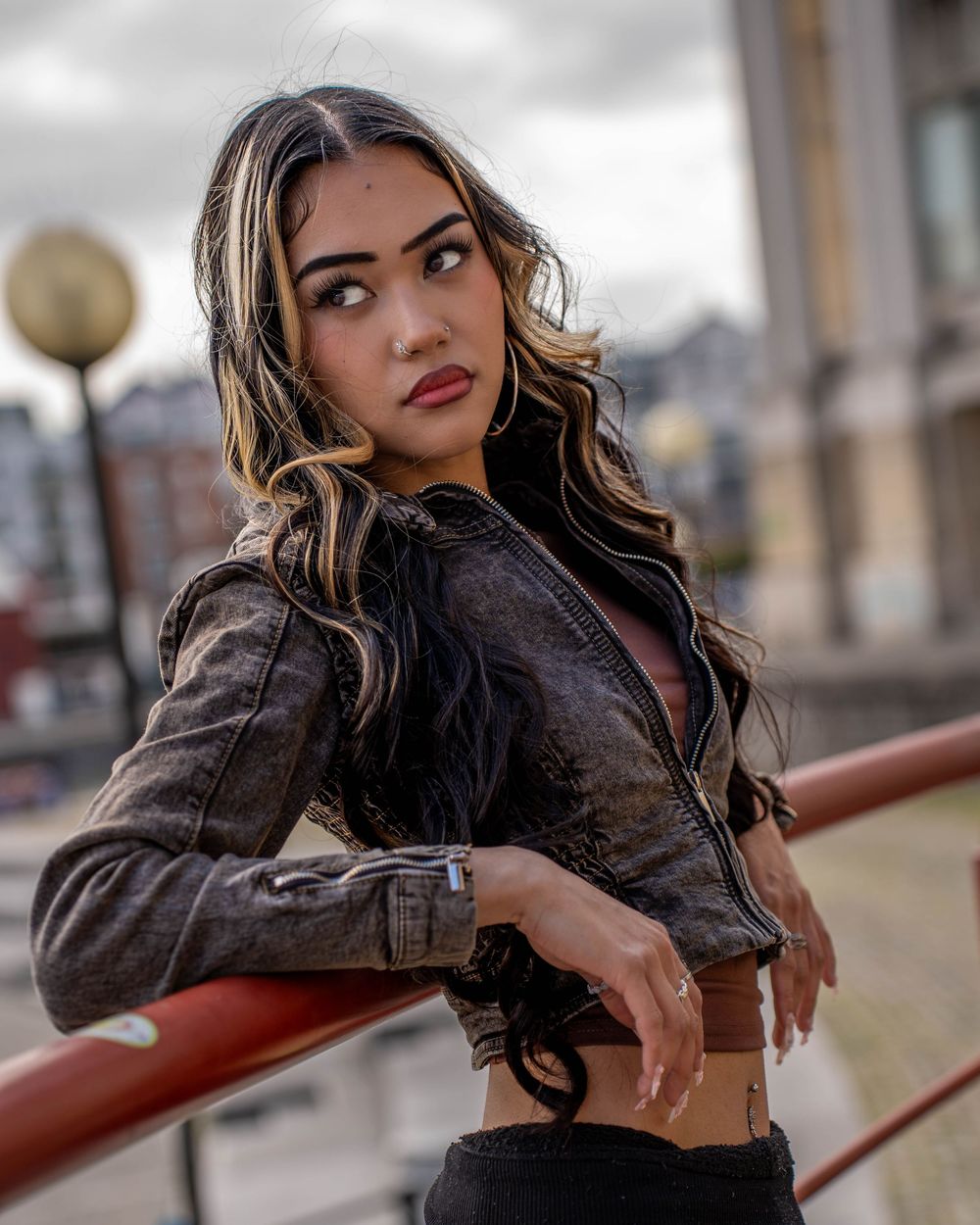 Outdoor fashion portrait with model in leather jacket leaning on red railing with urban background.