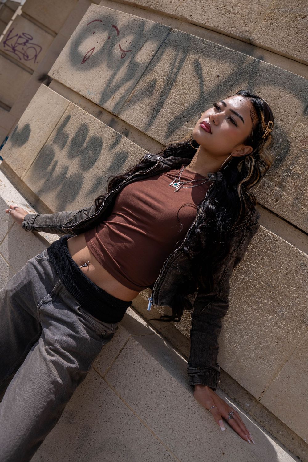 Street style photo of a model in brown crop top and black jacket posed against graffiti wall.