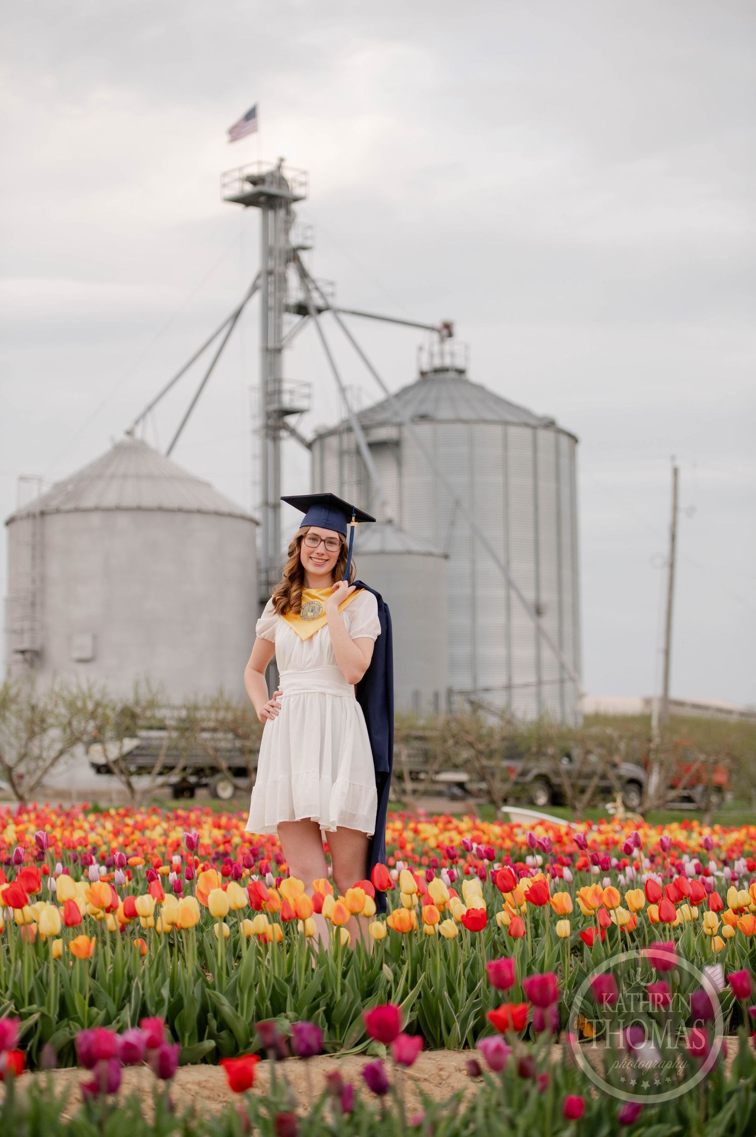 A graduate stands in a vibrant tulip field with colorful flowers blooming in front of large grain silos on an overcast day.