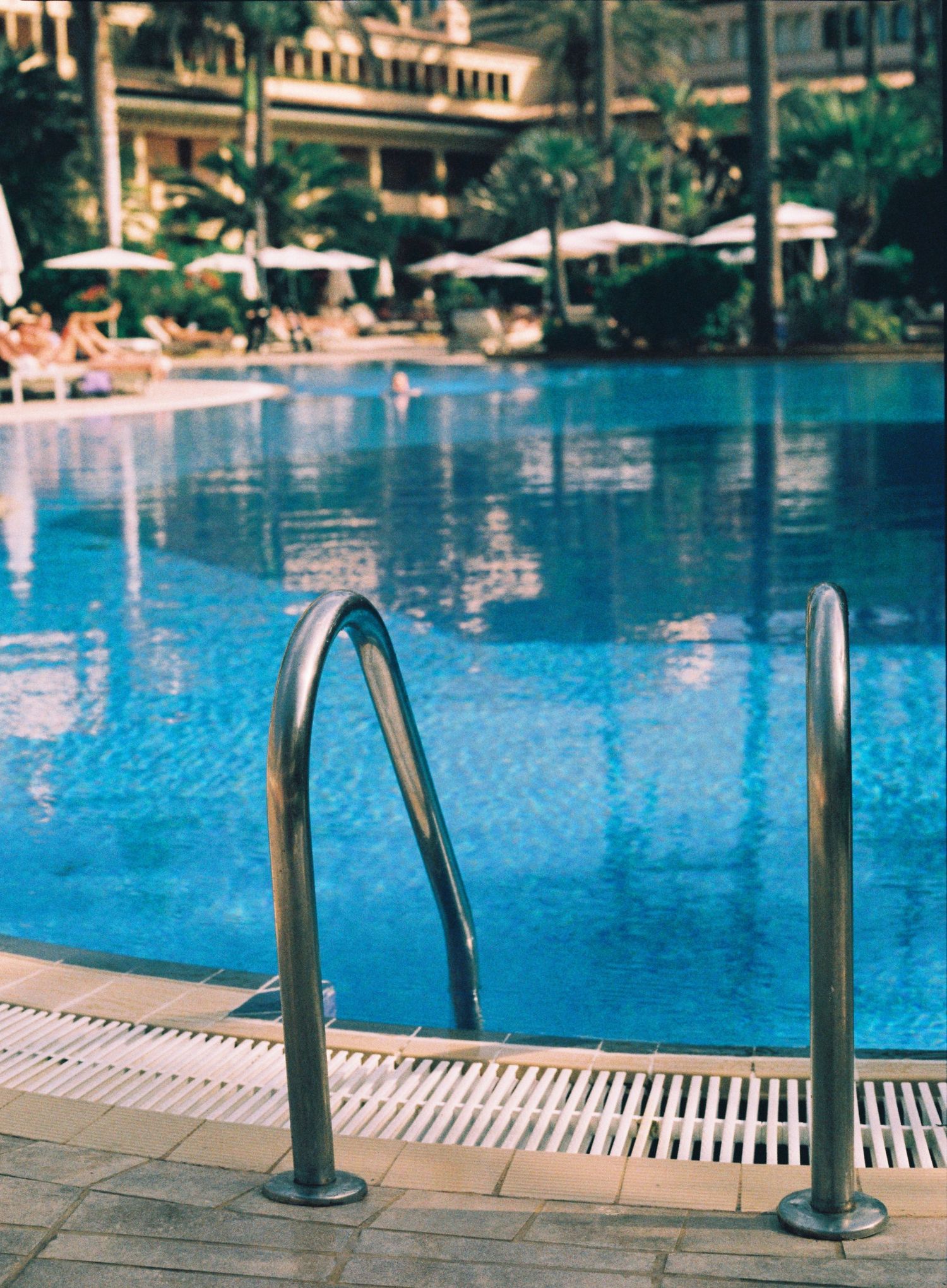 Stainless steel pool ladder leading into a luxurious blue swimming pool at a resort with palm trees in the background.