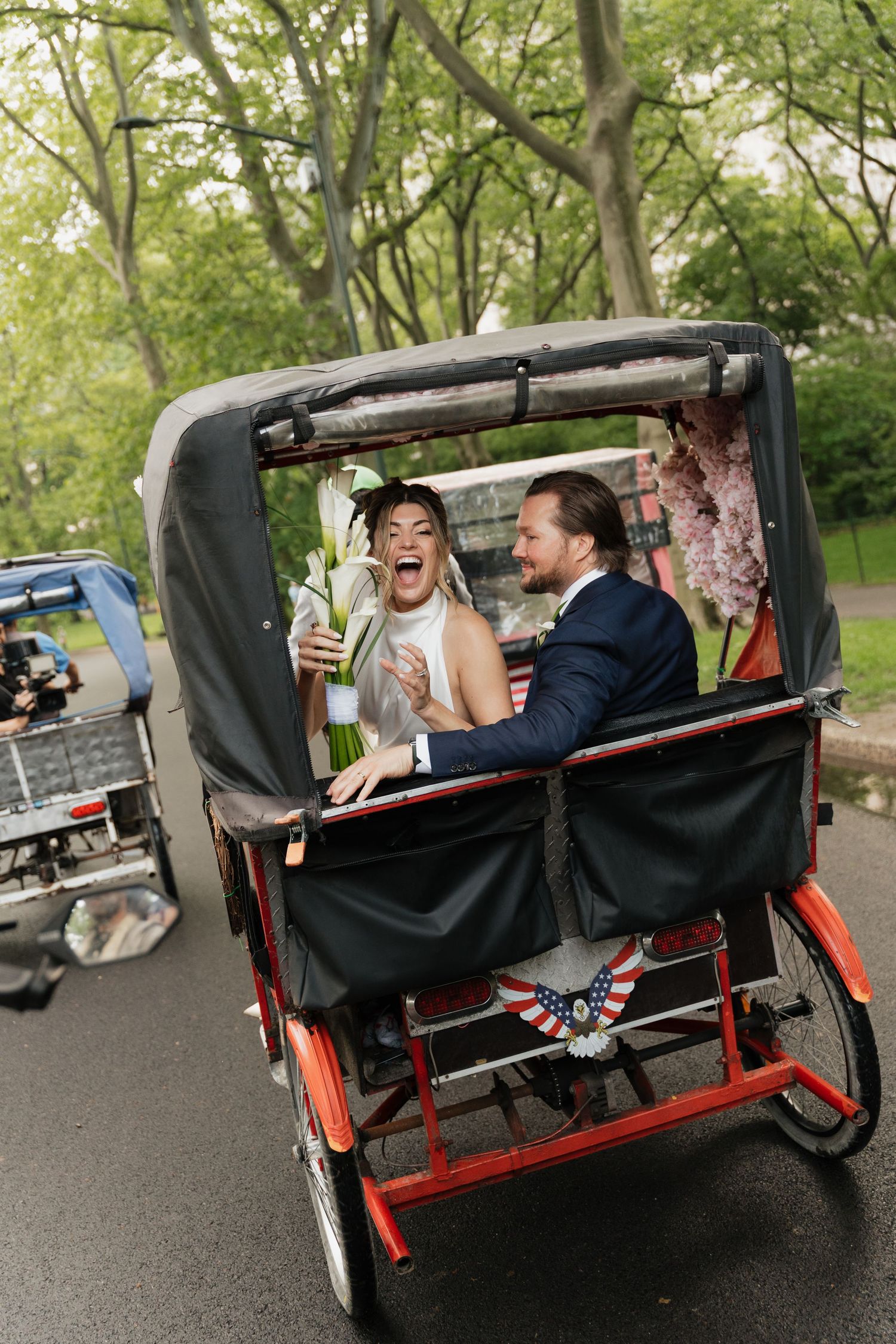 Two joyful passengers laugh while riding in a pedicab through a leafy park path on a sunny day.