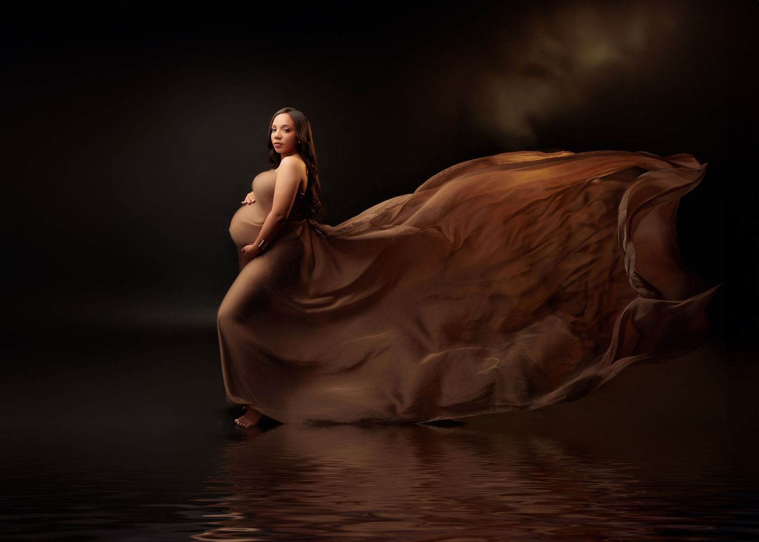 Pregnant woman draped in flowing brown fabric stands elegantly above reflective water on dark background.