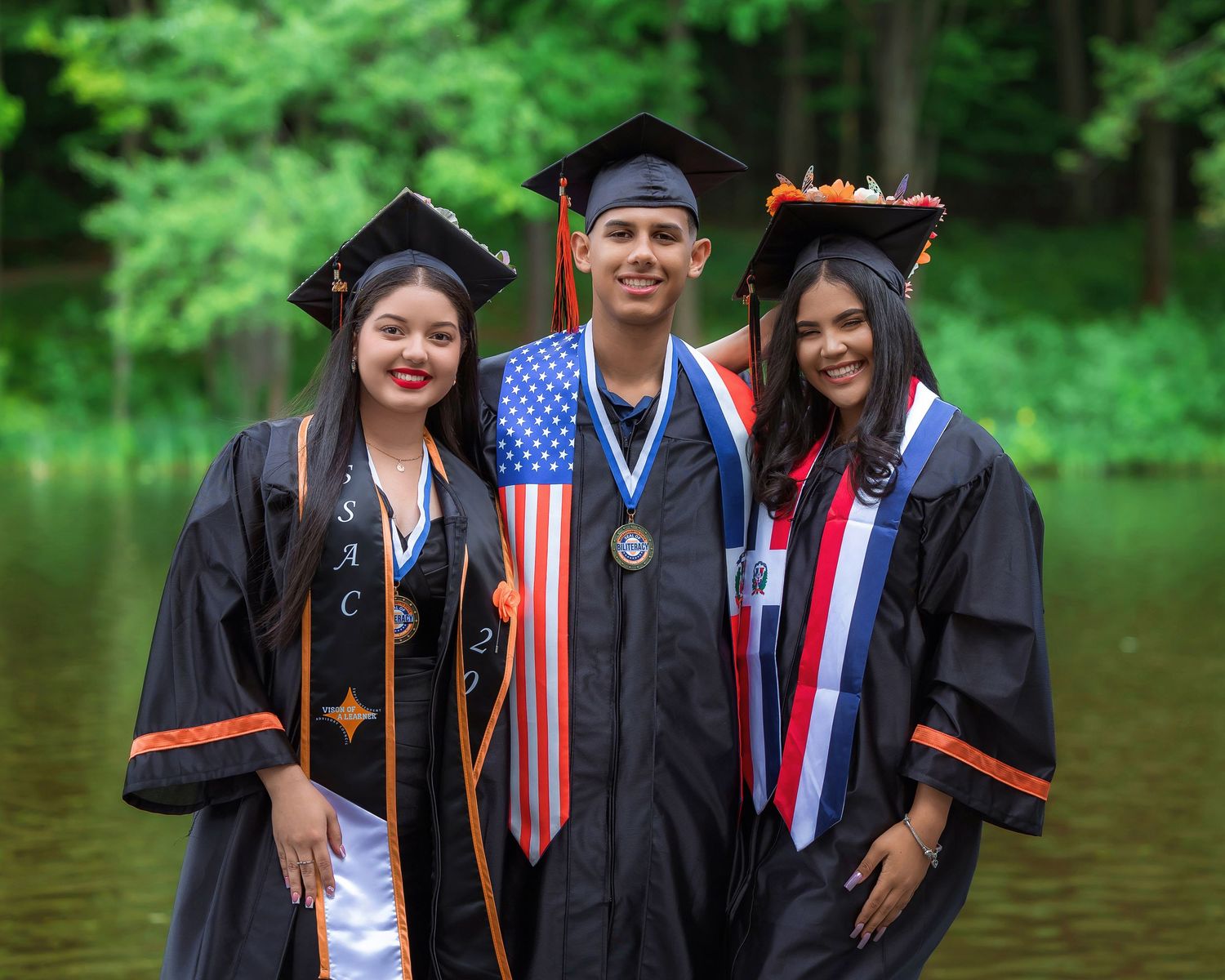 Three graduates in black caps and gowns posing outdoors near a green lake after their graduation ceremony.