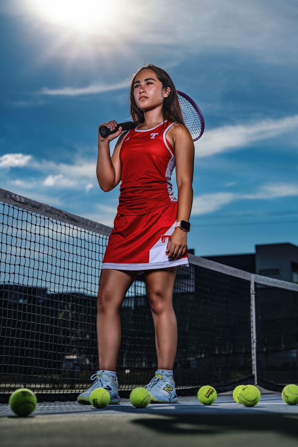 Tennis player in red uniform takes a break with tennis balls scattered on court under dramatic cloudy sky.