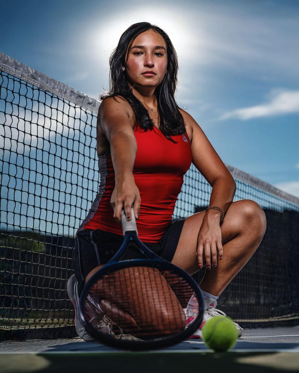 Athletic figure in red tank top and dark shorts poses with tennis ball on outdoor court in dramatic lighting.