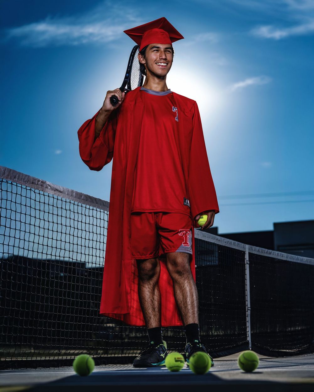 Graduate in red cap and gown poses on tennis court with balls scattered around during sunny outdoor photo session.