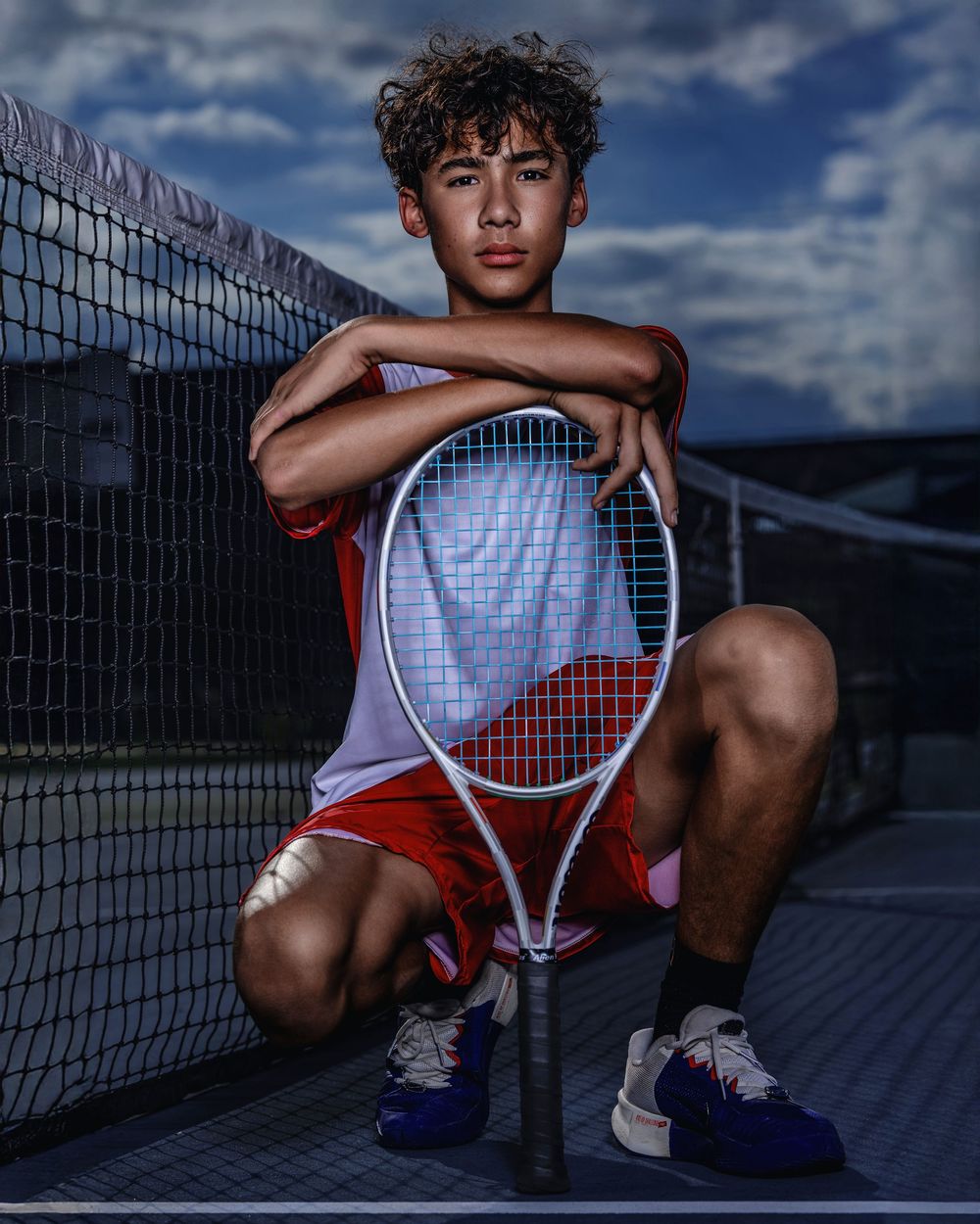 Young athlete poses with tennis racket against court fence at dusk wearing red shorts and white tank top.